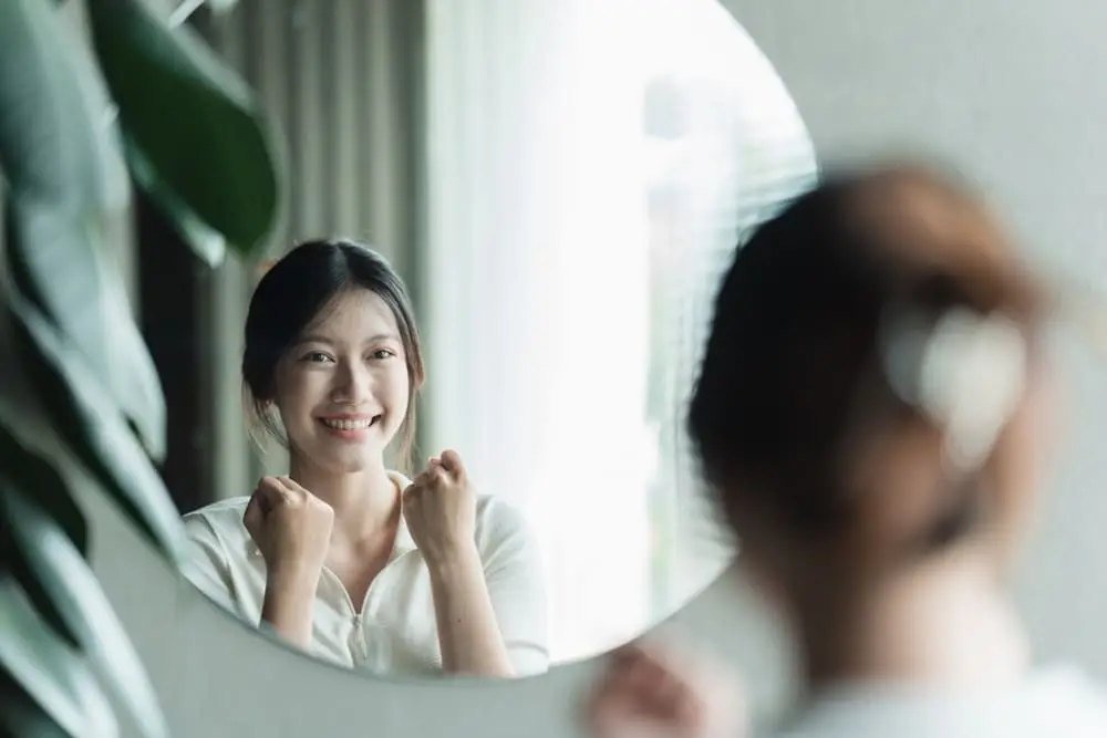 A woman standing in front of a mirror, smiling and practicing positive self-talk, reinforcing confidence and self-belief.